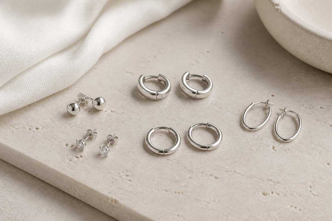 Small sterling silver stud earrings, huggie earrings, and slim hoops arranged on a light neutral background