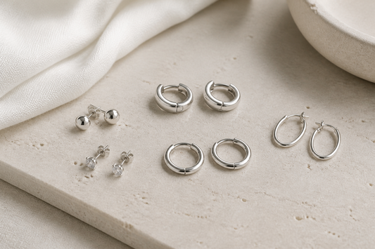 Small sterling silver stud earrings, huggie earrings, and slim hoops arranged on a light neutral background
